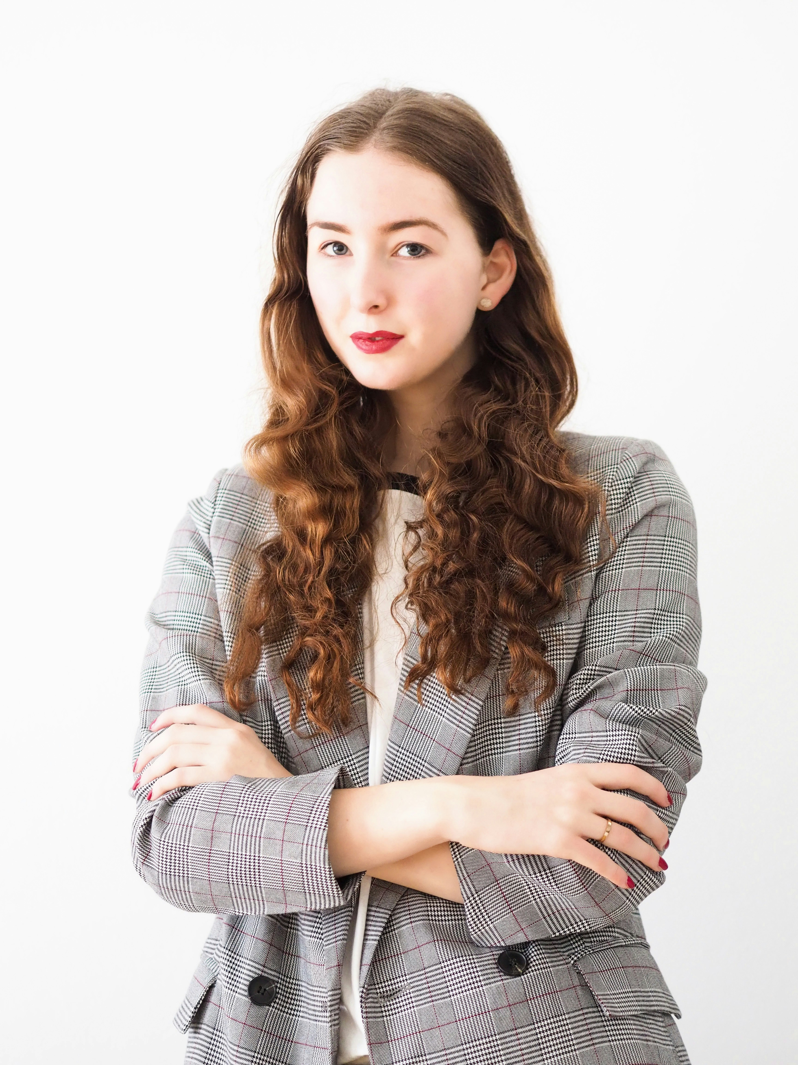 A woman with long, curly brown hair wearing a plaid blazer stands against a white background with her arms crossed.