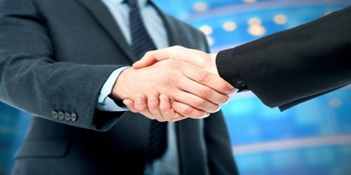 Two people in business attire shake hands against a blurred blue background, suggesting a professional agreement or partnership.