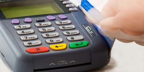 A hand swipes a credit card through a point-of-sale terminal with a numeric keypad and colored buttons.