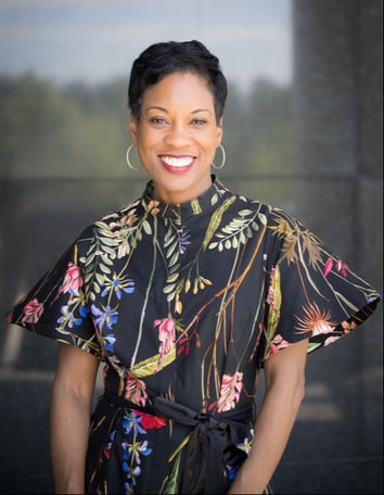 A woman stands smiling, wearing a floral-patterned black dress with short sleeves, hoop earrings, and a belt, in front of a blurred outdoor background.